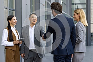 Adult males in business suits shaking hands while standing with caucasian women aside next to company office