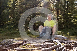 An adult man with a backpack sits in a sun-drenched clearing with tree roots