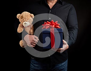 Adult man in a black shirt holds a blue square box tied with a red ribbon and brown teddy bear