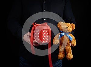 Adult man in a black shirt holds a blue square box tied with a red ribbon and brown teddy bear, concept of congratulations