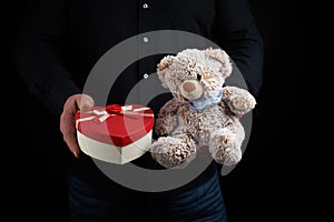 Adult man in a black shirt holds a blue square box tied with a red ribbon and brown teddy bear, concept of congratulations on