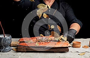 Adult man in a black shirt pours white coarse salt and a dry bay leaf on a fresh salmon filet