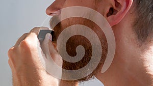 Adult man cutting his own beard and mustache with scissors and comb. Caucasian red bearded male trimming hair on face at home 4K