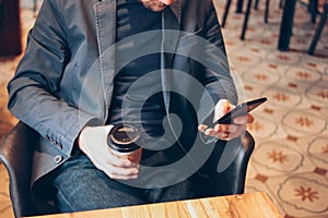 Adult man drinking coffee from paper cup and using mobile phone at cafe