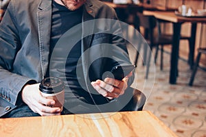 Adult man drinking coffee from paper cup and using mobile phone at cafe