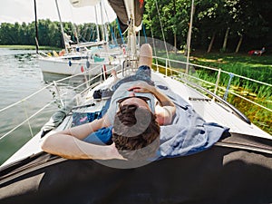 Adult man laying on a sailboat deck and taking sunbath during his vacations in a sunny summer day. Yacht moored on a wooden pier