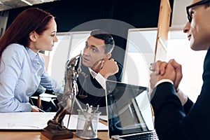 Adult man listens to red-haired woman at lawyer`s desk, where statue of Themis stands.