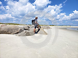 Adult man photographer in straw hat and shorts stands on ocean sand beach. In the hands of a camera behind the back there is a