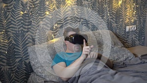 An adult man puts on virtual reality glasses and flips through virtual pages, lying on the bed in the bedroom in the
