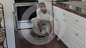 Adult man is putting round robotic vacuum cleaner on floor of kitchen, switching