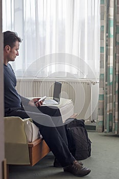 Adult man reads news on phone and laptop in hotel room