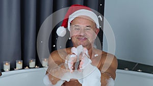 Adult man in santa claus hat sits in a big round bathtub, plays with foam, has fun, imitation of winter snow, christmas concept,