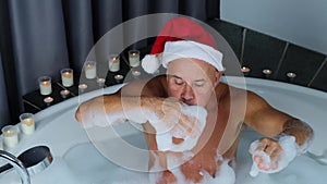 Adult man in santa claus hat sits in a big round bathtub, plays with foam, has fun, imitation of winter snow, christmas concept,