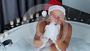 Adult man in santa claus hat sits in a big round bathtub, plays with foam, has fun, imitation of winter snow, christmas concept,