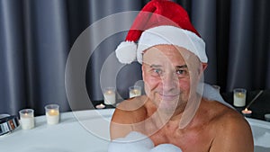 Adult man in santa claus hat sits in a big round bathtub, plays with foam, has fun, imitation of winter snow, christmas concept,