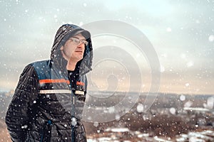 Adult man in snowy evening. Adult male in hooded outwear and glasses standing under falling snow against sunset window