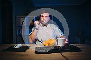 An adult man in a white T-shirt works at night at the computer and eats chips and tea sitting in a dark room with blue light