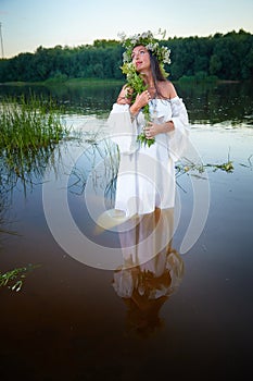 Adult mature brunette woman in a white dress, sundress and a wreath of flowers in summer in water of river or lake in