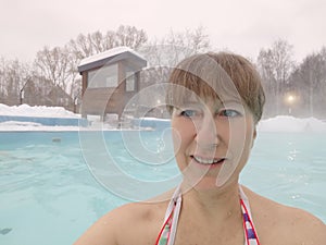 Adult mature woman takes a selfie in a pool with warm hot termal mineral water in winter and white snow around. Wellness