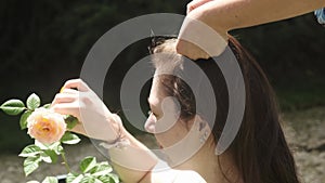 An adult mother styles her young daughter's long hair with her hands in an orchard next to a rose bush.