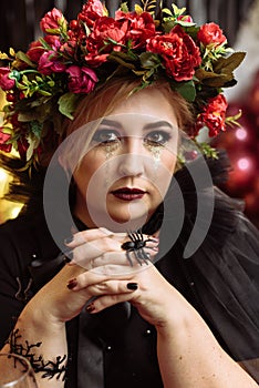 An adult plump woman in the image of a witch with flowers on her head sits in a dark studio