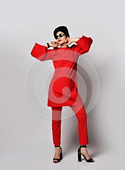 Adult pretty sensual short haired brunette woman is posing in stylish casual red costume tunic, pants, cap, sunglasses