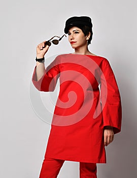 Adult pretty sensual short haired brunette woman in stylish casual red costume tunic, pants, cap puts on her sunglasses