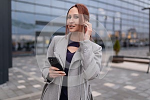 adult red-haired businesswoman speaks on a mobile phone through headphones on the background of a business center
