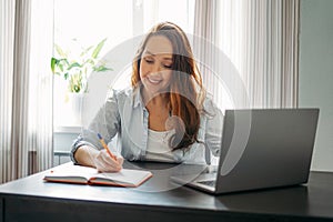 Adult smiling brunette woman doing notes in daily book with opened laptop. She study something with online course at the home