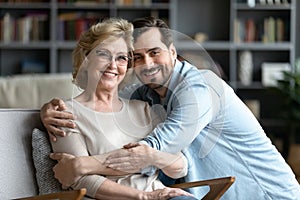 Adult son hugs elderly mother while she resting on armchair