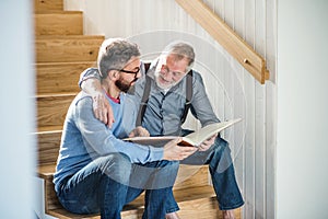 An adult son and senior father sitting on stairs indoors at home, looking at photographs.