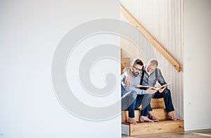 An adult son and senior father sitting on stairs indoors at home, looking at photographs.