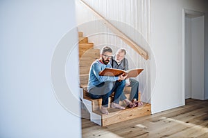 An adult son and senior father sitting on stairs indoors at home, looking at photographs.