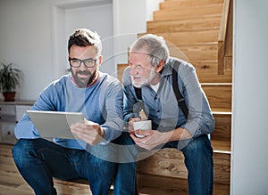 An adult son and senior father with tablet sitting on stairs indoors at home.