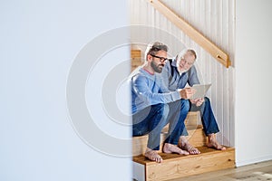 An adult son and senior father with tablet sitting on stairs indoors at home.