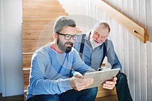 An adult son and senior father with tablet sitting on stairs indoors at home.