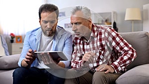 Adult son and senior father watching tutorial videos on tablet, information