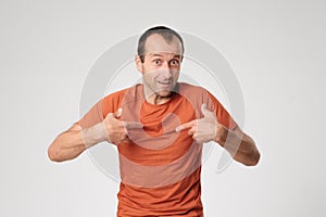 An adult Spanish man in an orange T-shirt points to himself with his finger.