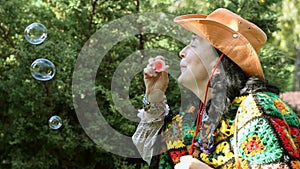 Adult stylish woman in a cowboy hat and a multicolored poncho blows bubbles.