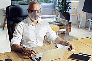 Adult successful businessman wearing a classic glasses and working at the wood table in modern coworking studio.Stylish