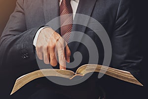 A man in a suit pointing a text on an open bible on a black background