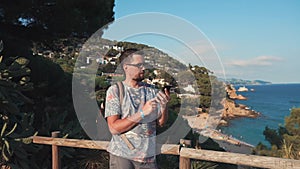 Adult townsman is standing in background of sea coastline, using smartphone
