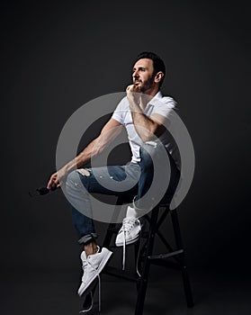 Adult unshaved man in white polo shirt, modern torn jeans and sneakers sits on high chair, stepladder looking aside