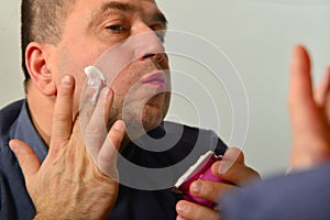 An adult and unshaven man in a blue shirt applies a mask of cream on his face, the concept of fashion, style and personal care