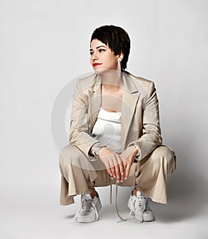 Adult woman in beige business casual pantsuit and sneakers sits squatted looking aside. Stylish business female wear