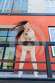 Adult woman in beige suit stands near brick building and holds reusable thermo mug in her hands