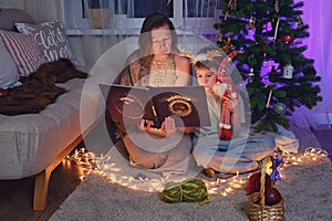 An adult woman and a boy are reading a book by the Christmas tree in