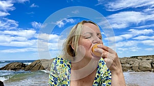 an adult woman in a bright dress on the beach greedily eats an orange, she bites it whole, pleasure is expressed on her