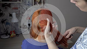 An adult woman combs the beautiful red hair of her teenage daughter, who is sitting in front of her on a chair in the