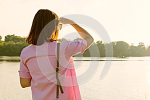 Adult woman enjoying summer sunset standing on the bank of the river, beautiful summer landscape, woman`s back looking into the d